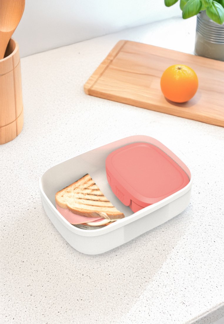 Biała śniadaniówka na blacie z kanapką i różowym pojemnikiem; pomarańcza na drewnianej desce do krojenia i zielona roślina w tle.