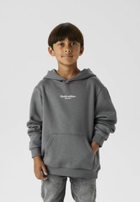 Grijze hoodie met een voorzak en witte tekst "Studio editie". De stof lijkt zacht; heeft een relaxed fit en ribgebreide manchetten.