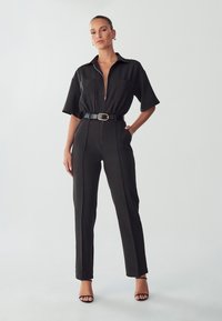 Zwarte jumpsuit met een ritssluiting aan de voorkant, korte mouwen, twee voorzakken en een ceintuur om de taille. Gladde stof met zakelijke rechte pijpen.