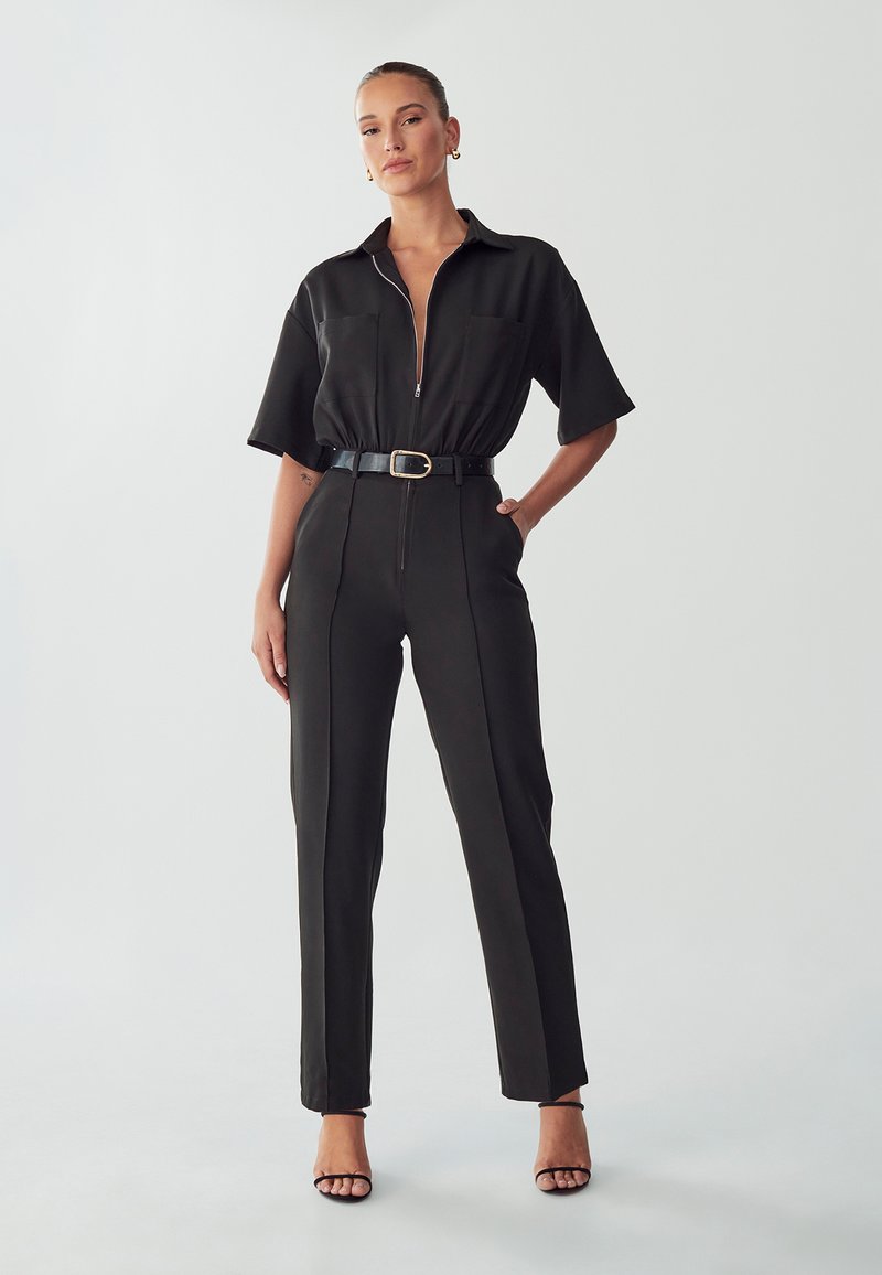 Zwarte jumpsuit met een ritssluiting aan de voorkant, korte mouwen, twee voorzakken en een ceintuur om de taille. Gladde stof met zakelijke rechte pijpen.