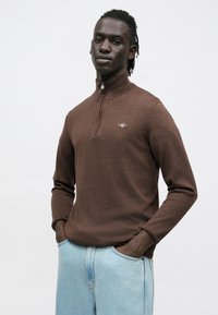 Brūns džemperis ar pusapkakli un rāvējslēdzēju, izgatavots no mīksta auduma. Uz krūtīm ir smalks logotips, un tam ir garas piedurknes.