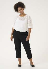 Haut blanc à manches bouffantes avec un motif de maille hexagonale, associé à un pantalon noir court. Les chaussures sont des ballerines noires. Silhouette simple et épurée.