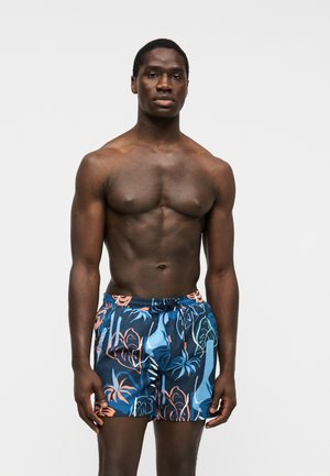 Uomo muscoloso in piedi fermo che indossa pantaloncini da bagno blu navy con motivi di foglie tropicali arancioni e azzurre su uno sfondo semplice.