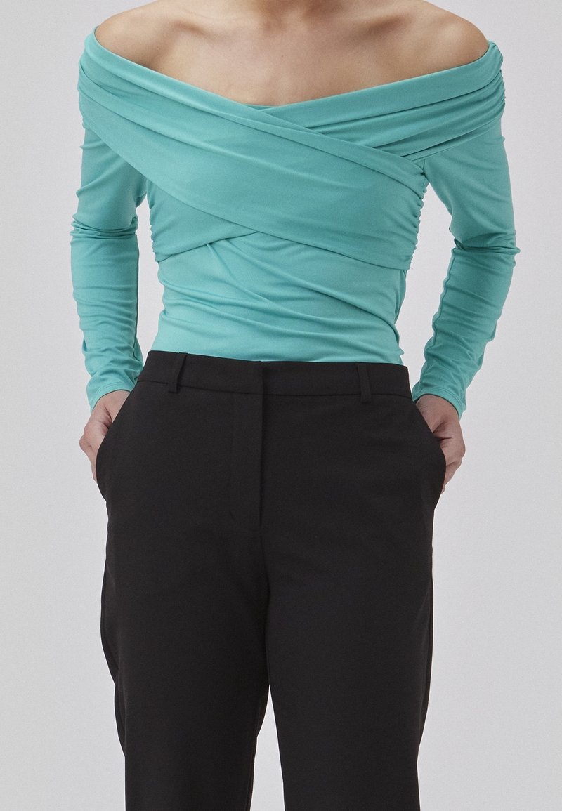 Teal off-the-shoulder top med lange ærmer, der har samlet stof hen over fronten, båret med sorte bukser. Glat, strækmateriale.
