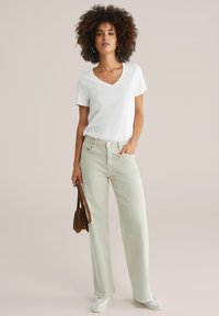 Bílý tričko s výstřihem do V je spárováno se světlými zelenými širokými džíny, doplněné hnědou crossbody taškou a světlými botami. Hladká textura látky.