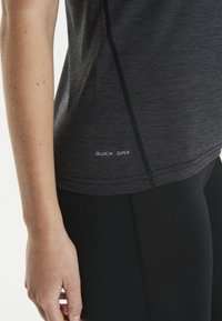 Sort hurtigttørrende træningsshirt med en meleret tekstur og flade sømme, nær en hånd på sorte leggings med lodrette sømdetaljer.