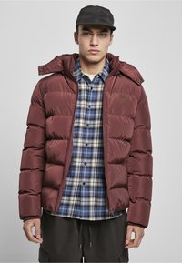 Chaqueta acolchada burdeos con capucha, que presenta un diseño acolchado. Lleva encima una camisa de franela a cuadros azules. Una gorra oscura completa el conjunto.