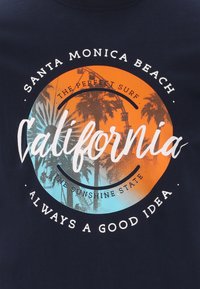 T-paita tummansinisellä pohjalla, jossa on pyöreä graafinen kuva oranssista ja sinisestä gradientista, palmun siluetteja sekä valkoista tekstiä Kaliforniasta.