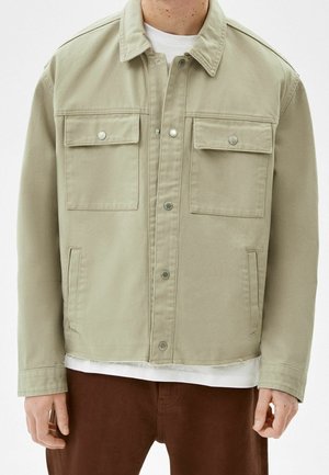 Beige Jeansjacke mit Knopfverschluss, zwei Brustklappentaschen und zwei seitlichen Schlitztaschen, getragen über einem weißen Hemd und braunen Hosen.