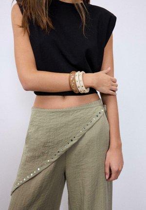 Ženska, ki nosi črno brezrokavnik brez rokavov in olivno zelene hlače z visokim pasom ter okrasnimi okroglimi zakovicami ob sprednji preklopni zanki.