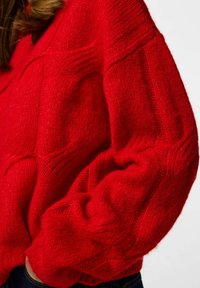Czerwony sweter z dzianiny o miękkiej fakturze, oversize'owym kroju i prążkowanych wykończeniach na mankietach i dole. Wykończony wzorzystym detalem.