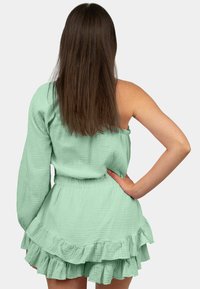 Mintgroene jurk met één lange mouw, losse pasvorm, textuurstof, gerimpelde zoom en elastische tailleband, gezien van achteren.