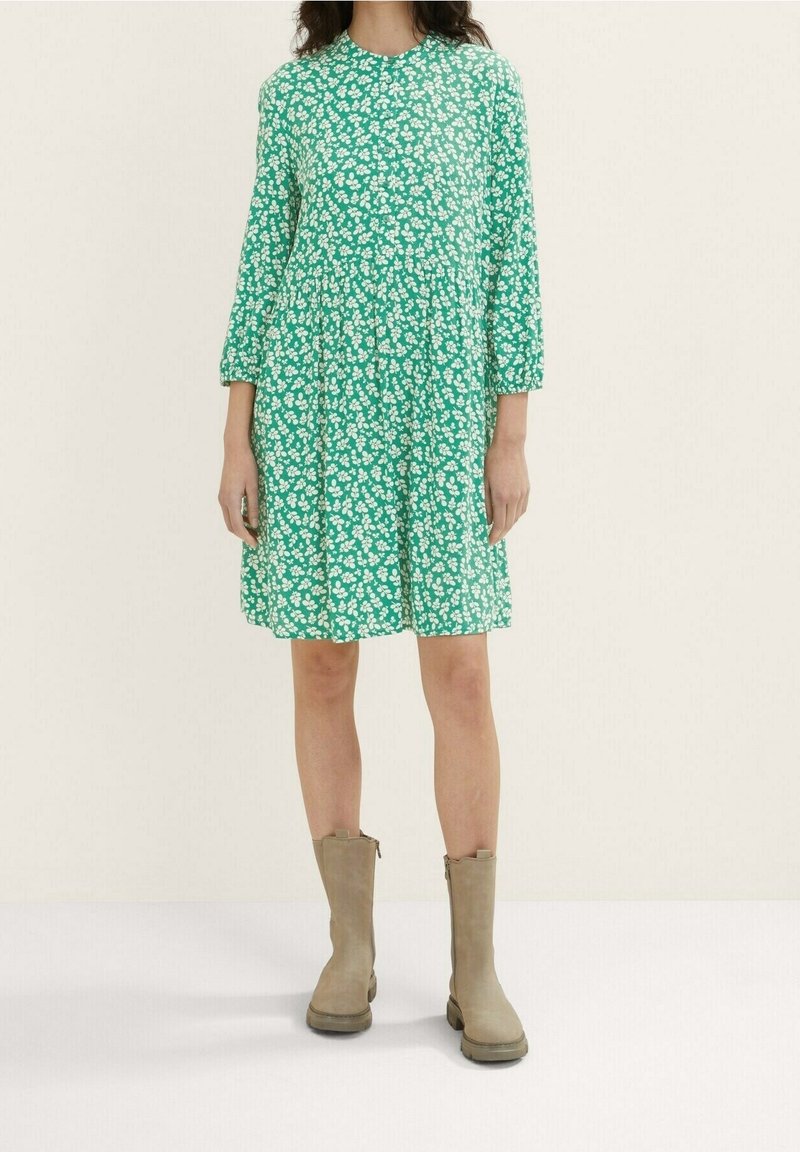 Femme portant une robe verte à fleurs mi-longue avec manches longues et des bottes beige mi-mollet, debout devant un fond clair uni.