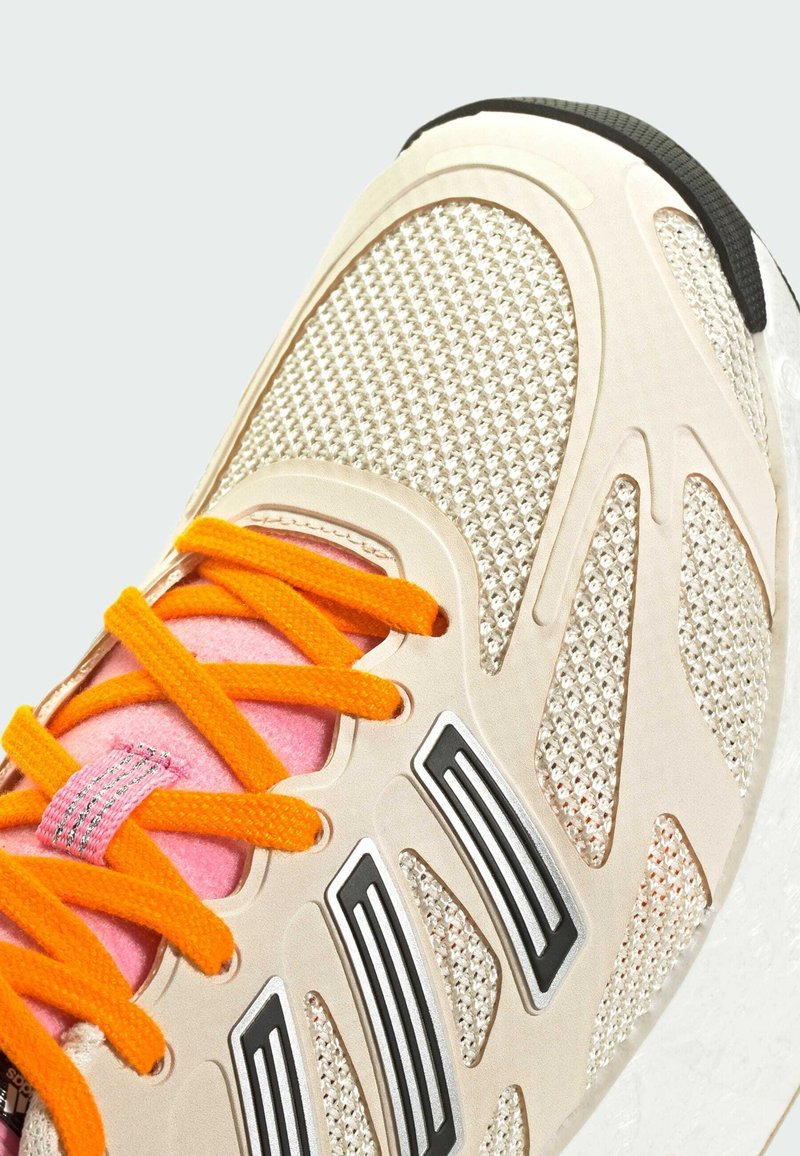 Sneaker mit einem hellbeigen Mesh-Obermaterial, orangen Schnürsenkeln, pinken Akzenten und schwarzer Sohle. Verfügt über geometrische Aussparungen und metallische Details.