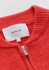 Rode gebreide trui met een geribbelde halslijn, voorzien van een zilveren rits. Etiket leest "OPUS," maat 36, op een wit label. Zachte textuur.