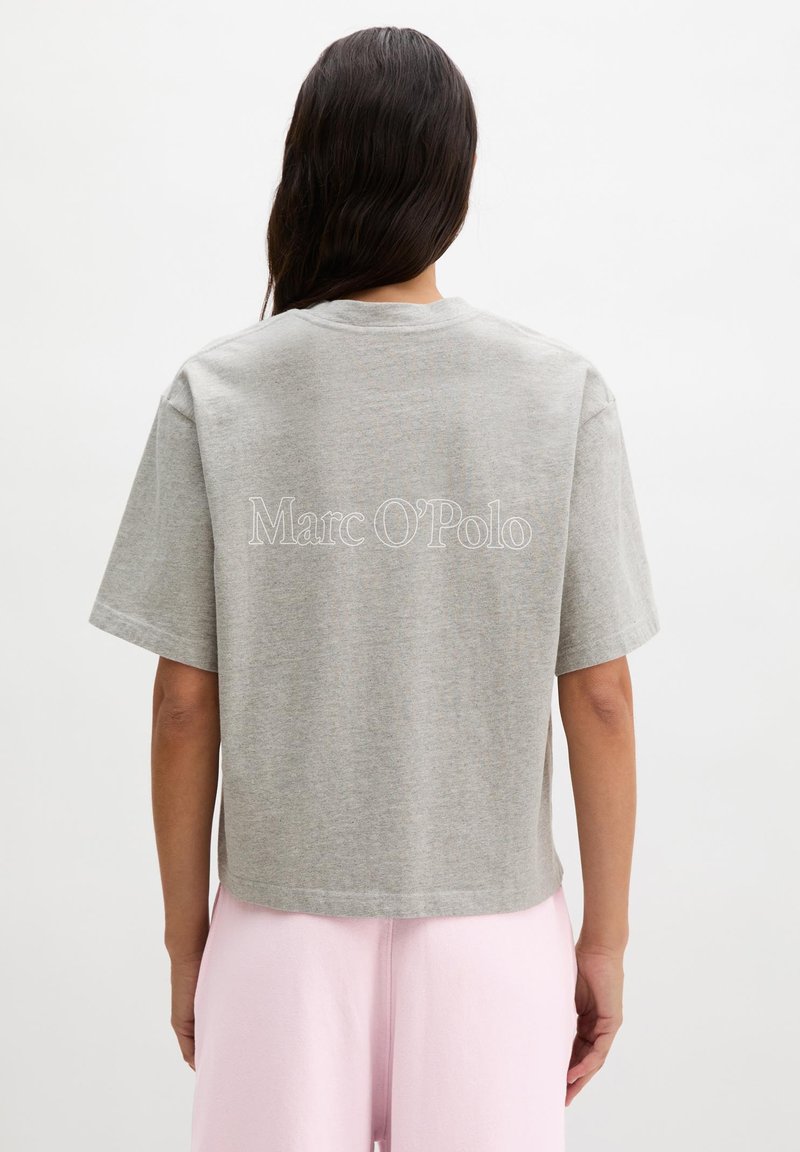 Femme aux longs cheveux foncés portant un T-shirt gris Marc O'Polo et un pantalon rose clair, debout de dos devant un fond uni.