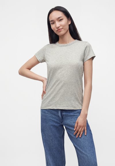 Femme aux longs cheveux foncés portant un t-shirt gris clair à manches courtes et un jean bleu, debout avec une main sur la hanche.