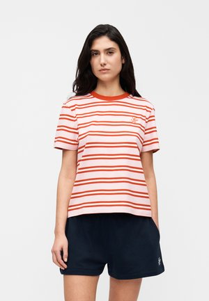 Woman with dark hair wearing a pink and red striped t-shirt and navy blue shorts standing against a plain white background.