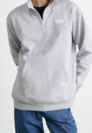 Osoba ubrana w jasnoszarą bluzę North Face z półzamkiem i przednią kieszenią oraz niebieskie dżinsy, stoi z rękami za plecami.