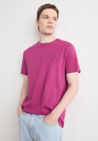 Magenta trumpas rankovėmis marškinėliai iš medvilnės, su apvaliu kaklu ir apvaliu logotipu kairėje krūtinės pusėje. Atsitiktinis siluetas.