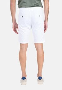 Pantaloni corto in cotone bianchi con due tasche posteriori e bottoni neri, caratterizzati da un taglio dritto e una rifinitura pulita all'orlo. Un paio di scarpe casual visibili.