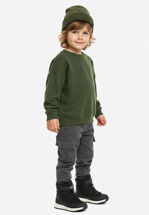Niño pequeño con gorro verde y suéter, pantalones cargo grises y botas de invierno negras, de pie sobre un fondo blanco.