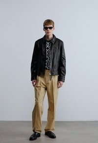 Μαύρο δερμάτινο μπουφάν, μαύρο πουκάμισο με σχέδια, μπεζ παντελόνια και μαύρα παπούτσια. Το μοντέλο φοράει σκούρα γυαλιά ηλίου. Απλό φόντο.