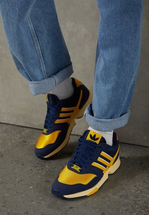 Person trägt marineblaue und gelbe Sneakers mit weißen Socken und hochgekrempelten blauen Jeans und steht auf einem grauen Betonboden.