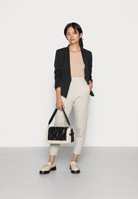 Schwarzes Blazer, beigefarbenes geripptes Oberteil, cremefarbene Hose und beigem Loafer. Hält eine schwarz-creamfarbene gesteppte Handtasche mit Schultergurt.