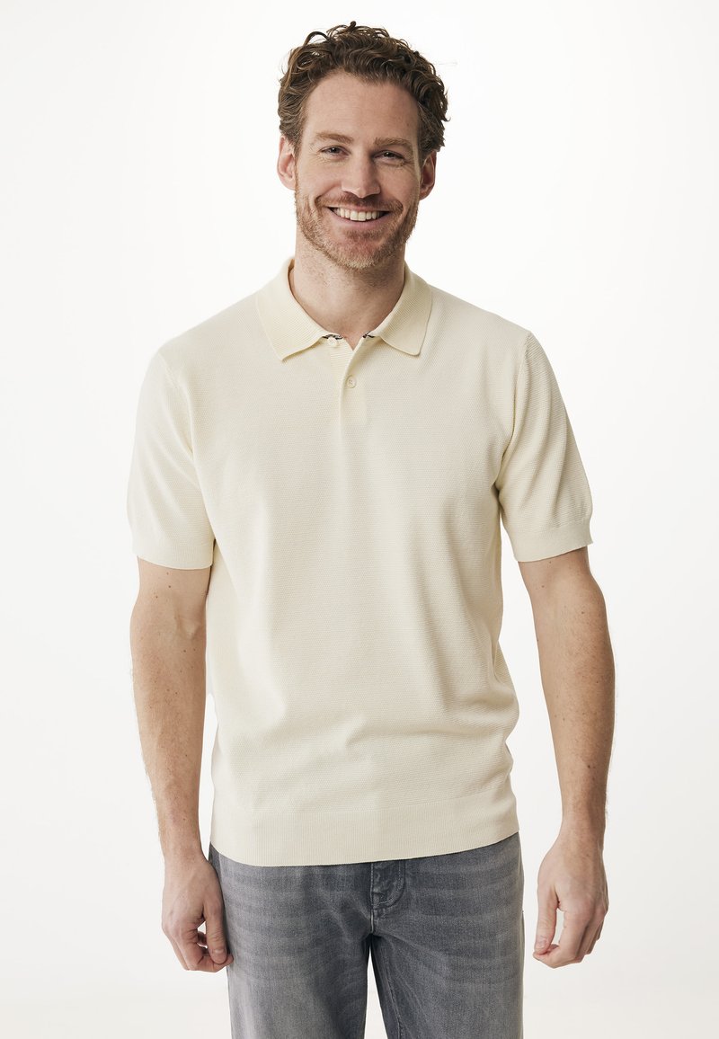 Pikétröja i mjukt krämfärgat tyg, med klassisk krage, korta ärmar och ett strukturerat mönster, parat med grå jeans.