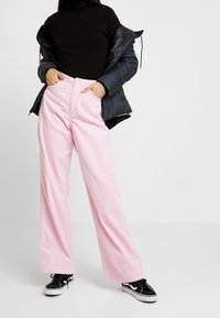 Pantalon large en velours côtelé rose avec rayures verticales, taille haute et boutons devant, associé à un col roulé noir ajusté et une veste noire bouffante.