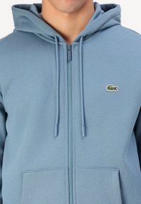 Haut à capuche zippé bleu clair en tissu doux, avec une poche kangourou, des cordons de serrage et un logo de crocodile brodé sur la poitrine.
