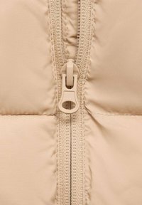 Detailný záber na béžovú páperovú bundu, obsahujúci hladkú štruktúru, horizontálne švy a zips s okrúhlym ťahákom.