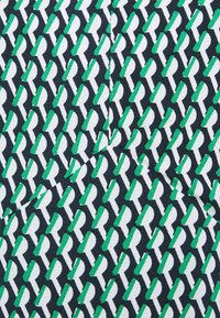 Tyg med ett upprepat geometriskt mönster av gröna, vita och svarta former mot en mörk bakgrund, med en slät textur.