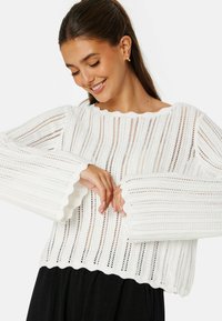 Weißer Strickpullover mit offenen Netzmustern, langen Glockenärmeln und einem gewellten Saum. Leichter und atmungsaktiver Stoff.