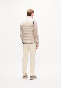 Bărbat stând de spate, purtând o vestă puffer bej, un pulover cu model, pantaloni crem și adidași bej, pe un fundal alb simplu.
