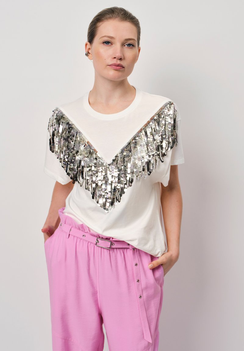 Witte t-shirt met een zilveren sequins v-vorm ontwerp over de borst. Gecombineerd met lichtroze broek met een verstelbare riem en drukknopen.