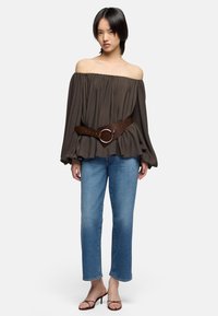 Femme portant un chemisier marron foncé à épaules dénudées avec des manches bouffantes, une large ceinture marron, un jean bleu clair et des sandales à talons marron.