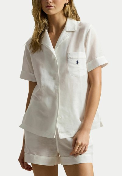 Vrouw draagt een witte, korte mouwen blouse met knopen en borstzak, gecombineerd met witte shorts, tegen een effen achtergrond.
