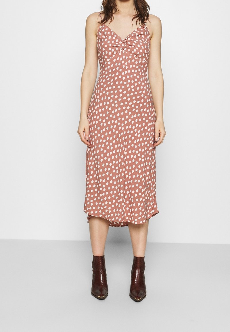 Robe midi rose à pois avec des bretelles fines, un détail froncé au niveau de la poitrine et une jupe fluide. Associée à des bottines noires en cuir.