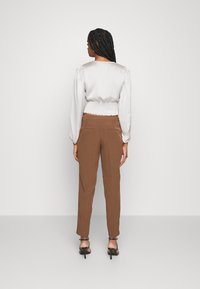 Blusa de seda cinza claro com mangas longas bufantes e cintura franzida, combinada com calças castanhas de perna estreita e sapatos de salto alto pretos.