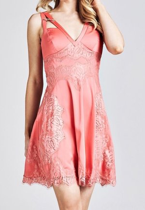 Kobieta ubrana w koralowo-różową sukienkę bez rękawów z koronkowymi zdobieniami i dekoracyjnym paskiem na jednym ramieniu, stojąca na jednolitym tle.