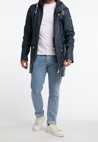 Veste imperméable bleu marine avec capuche, fermeture éclair à l'avant, deux poches avant, accents jaunes et détails à cordon. Portée par-dessus un t-shirt blanc et un jean.