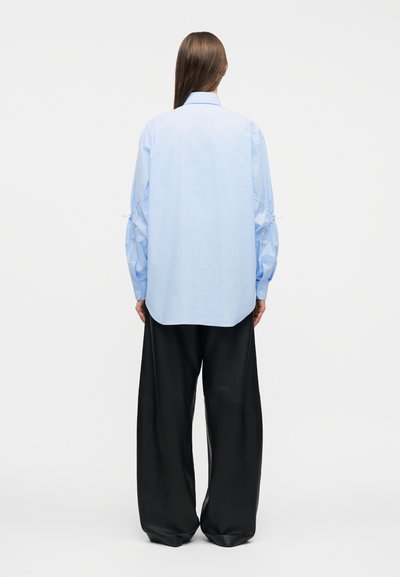 Άτομο που στέκεται γυρισμένο πίσω, φοράει ανοιχτό μπλε πουκάμισο με μακριά μανίκια με σούρες και φαρδιά μαύρη παντελόνα.