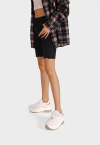 Sneakers bianchi con una tomaia testurizzata e accenti beige, abbinati a pantaloni shorts in denim neri e una camicia a quadri.