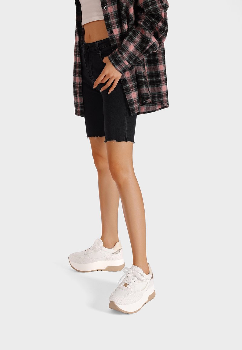 Sneakers bianchi con una tomaia testurizzata e accenti beige, abbinati a pantaloni shorts in denim neri e una camicia a quadri.