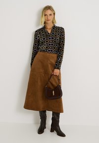 Jupe maxi en daim marron, blouse noire à manches longues à motifs, bottes en cuir marron foncé montantes, et un sac à main courbé avec des détails en or.