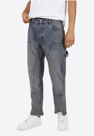 Persona che indossa pantaloni cargo in denim blu sbiadito con multiple tasche, una maglietta bianca e scarpe da ginnastica bianche, in piedi contro uno sfondo bianco.