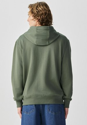 Mann mit lockigem Haar, der einen übergroßen olivgrünen Hoodie und blaue Jeans trägt, von hinten vor einem schlichten hellen Hintergrund.