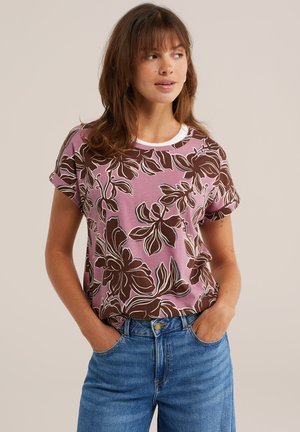 Frau trägt ein lila T-Shirt mit großem braunem Blumenmuster und blaue Jeans, steht mit den Händen in den Taschen und schaut zur Seite.